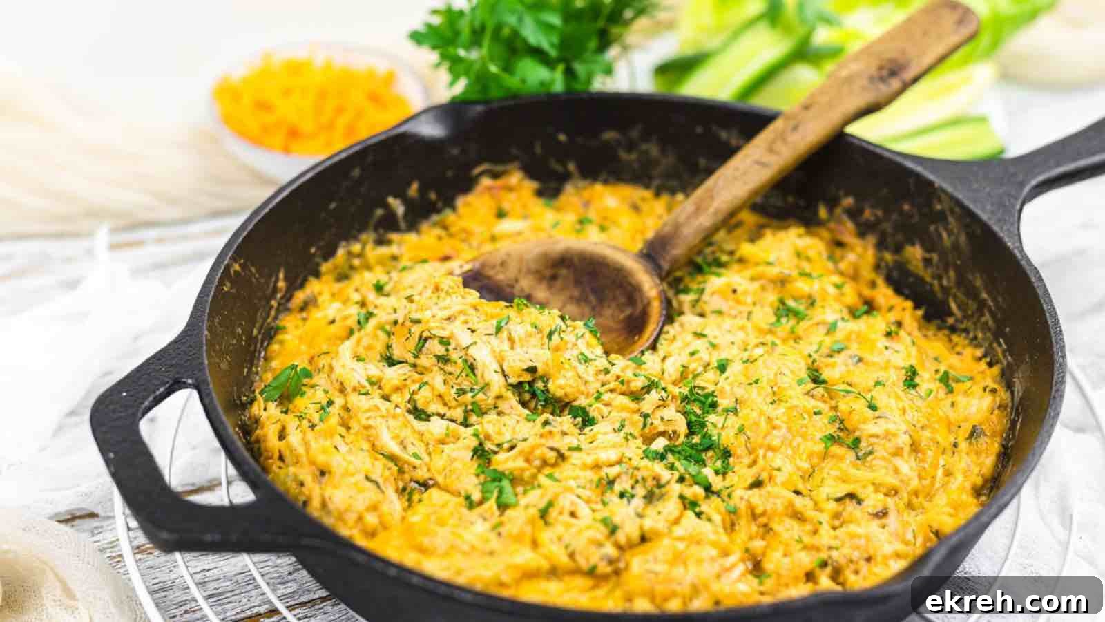 A cast iron skillet filled with a creamy dish, garnished with chopped herbs and a wooden spoon inside. Fresh parsley and sliced vegetables are seen in the background.