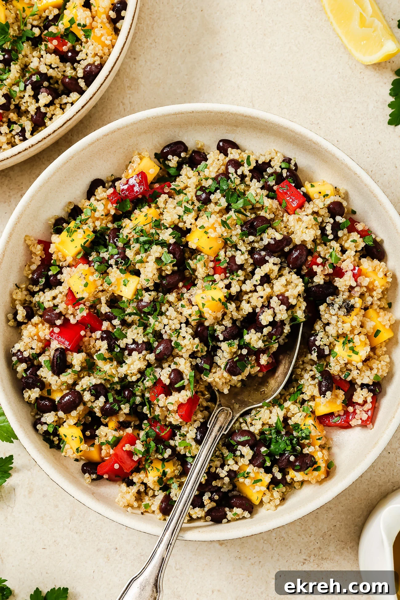 A bowl of mango quinoa salad with black beans, diced red and yellow bell peppers, chopped herbs, and a spoon, placed on a light surface with lemon wedges and parsley nearby.