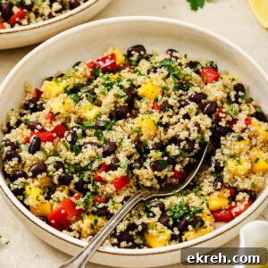 A vibrant mango quinoa salad featuring black beans, red bell peppers, yellow mango, and chopped parsley, served in a bowl with a spoon and a lemon wedge on the side.