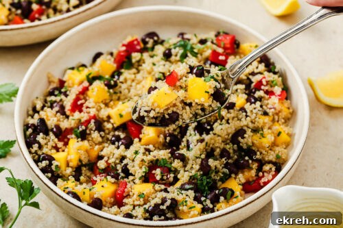 A close-up of a vibrant mango quinoa salad with black beans, diced mango, red bell peppers, and herbs. A spoon lifts a portion from the bowl, while lemon wedges and fresh parsley sit nearby.