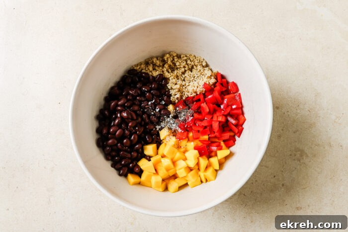 A white bowl contains separate portions of black beans, cooked quinoa, diced red bell pepper, diced mango, and seasoning on a light countertop—perfect for assembling a fresh mango quinoa salad.