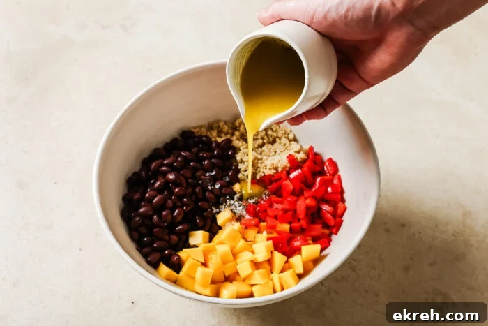 A hand pours yellow dressing from a small white jug into a vibrant mango quinoa salad, filled with black beans, chopped red bell pepper, and diced mango on a light countertop.