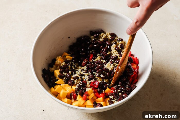 A hand stirs a bowl filled with black beans, quinoa, diced yellow mango, and red bell peppers—classic ingredients for a vibrant mango quinoa salad—using a wooden spoon on a light countertop.