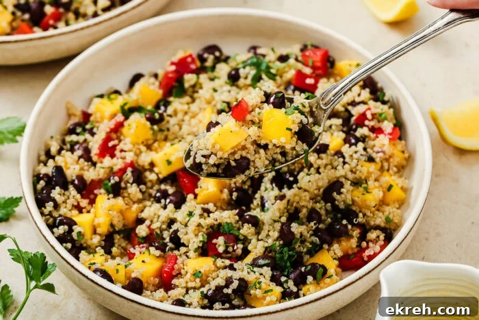 A close-up of a vibrant mango quinoa salad with black beans, diced mango, red bell peppers, and herbs. A spoon lifts a portion from the bowl, while lemon wedges and fresh parsley sit nearby.