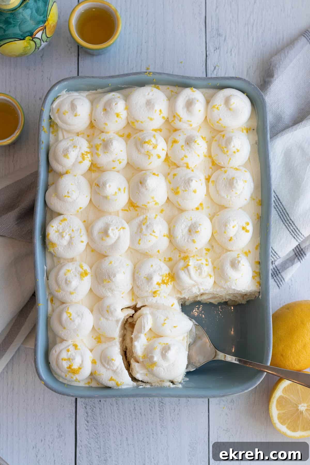 Limoncello Tiramisu Recipe with Zesty Lemon Mascarpone 5 blue baking dish of limoncello tiramisu with a spoon scooping out a serving and lemons and limoncello in the foreground and background