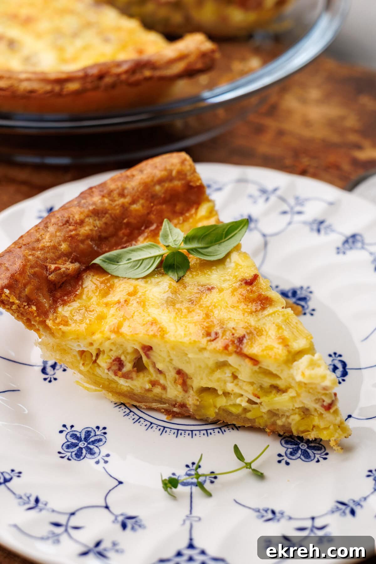 A slice of quiche lorraine on a vintage plate with the rest of the quiche in the background in a glass pie dish