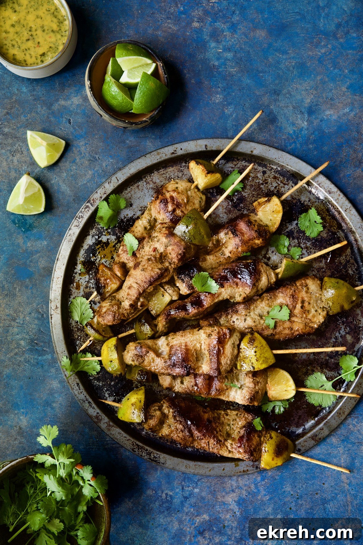 Grilled Citrus Mojo Pork Tenderloin Skewers 12 Mojo-Marinated Pork Skewers on metal tray with dipping sauce, lime wedges and cilantro leaves.