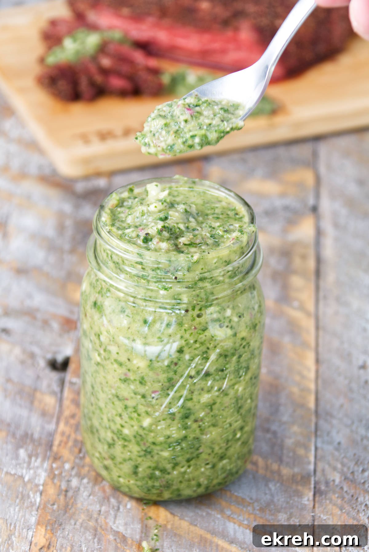 mason jar filled with vibrant green cilantro chimichurri sauce with sliced smoked tri-tip on the background on a cutting board