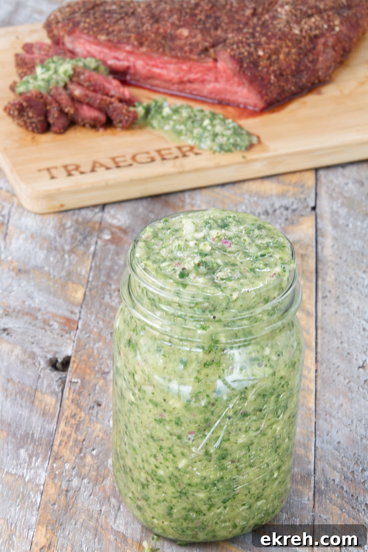 mason jar filled with vibrant green cilantro chimichurri sauce with sliced smoked tri-tip on the background on a cutting board