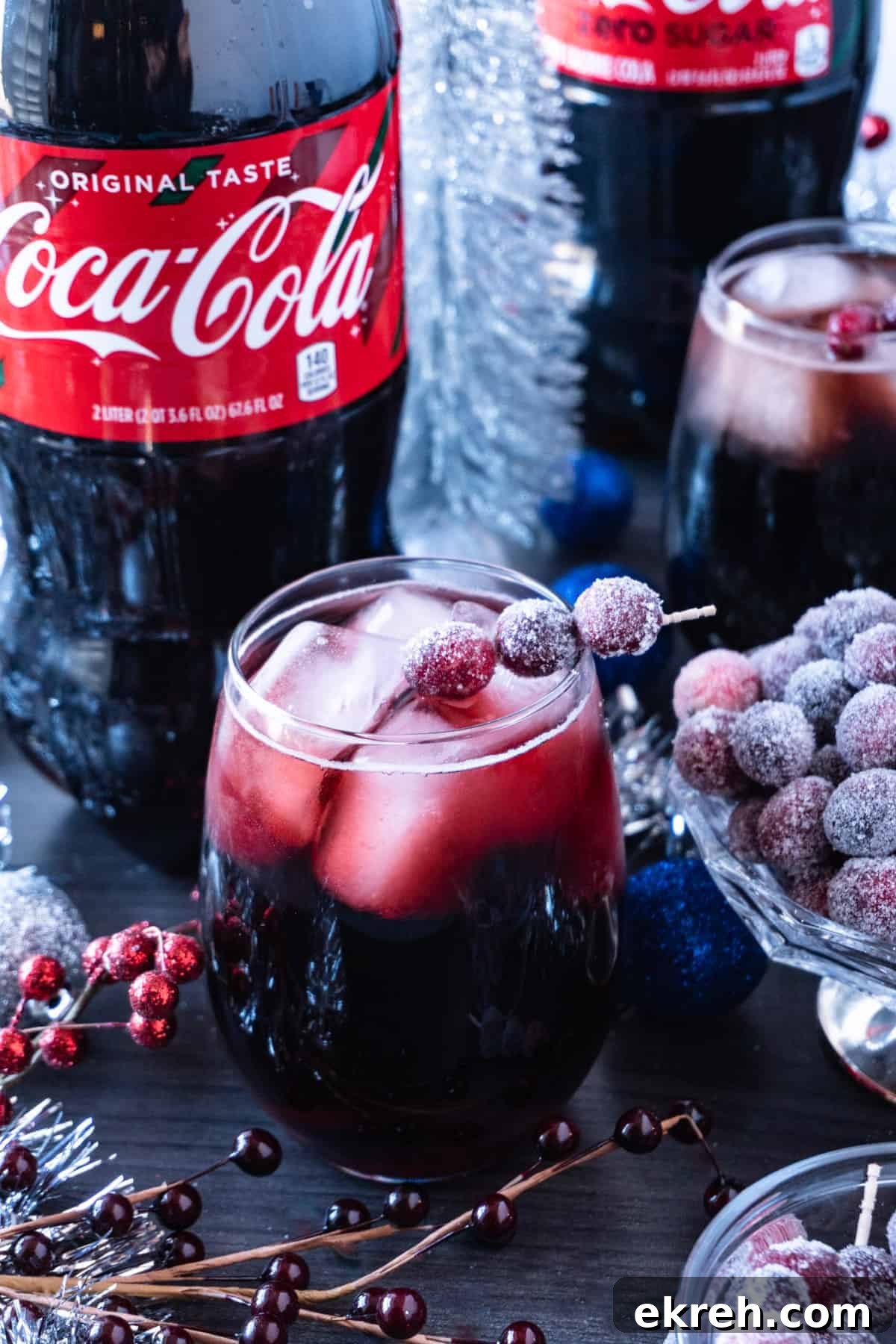 Quick Christmas Cocktail Recipe 4 Christmas drink in a clear glass with large ice cubes and a toothpick with sugared cranberries on it to garnish the drink with a 2L bottle of Coca-Cola in the background and Christmas decorations all around the drink.