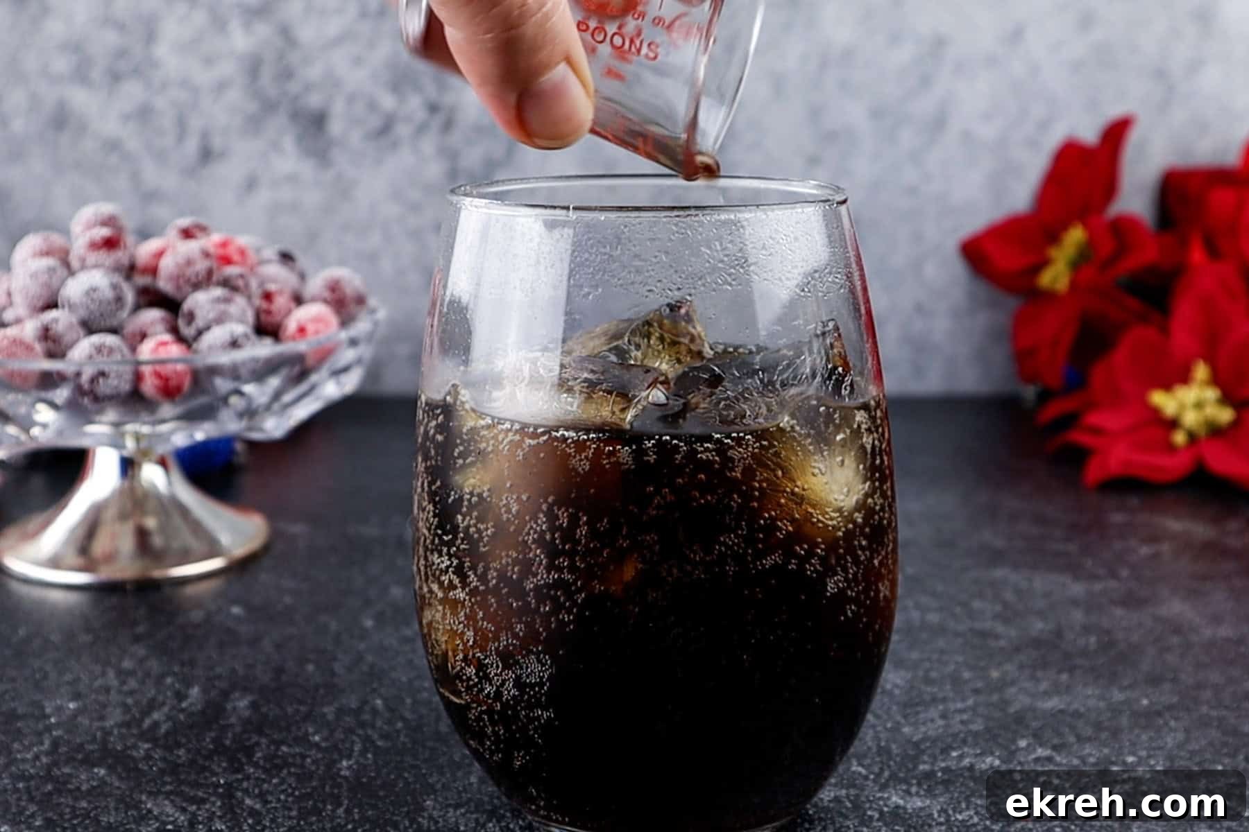 Quick Christmas Cocktail Recipe 6 Frosted glass of soda with ice cubes, pouring vanilla into glass, festive holiday setting.
