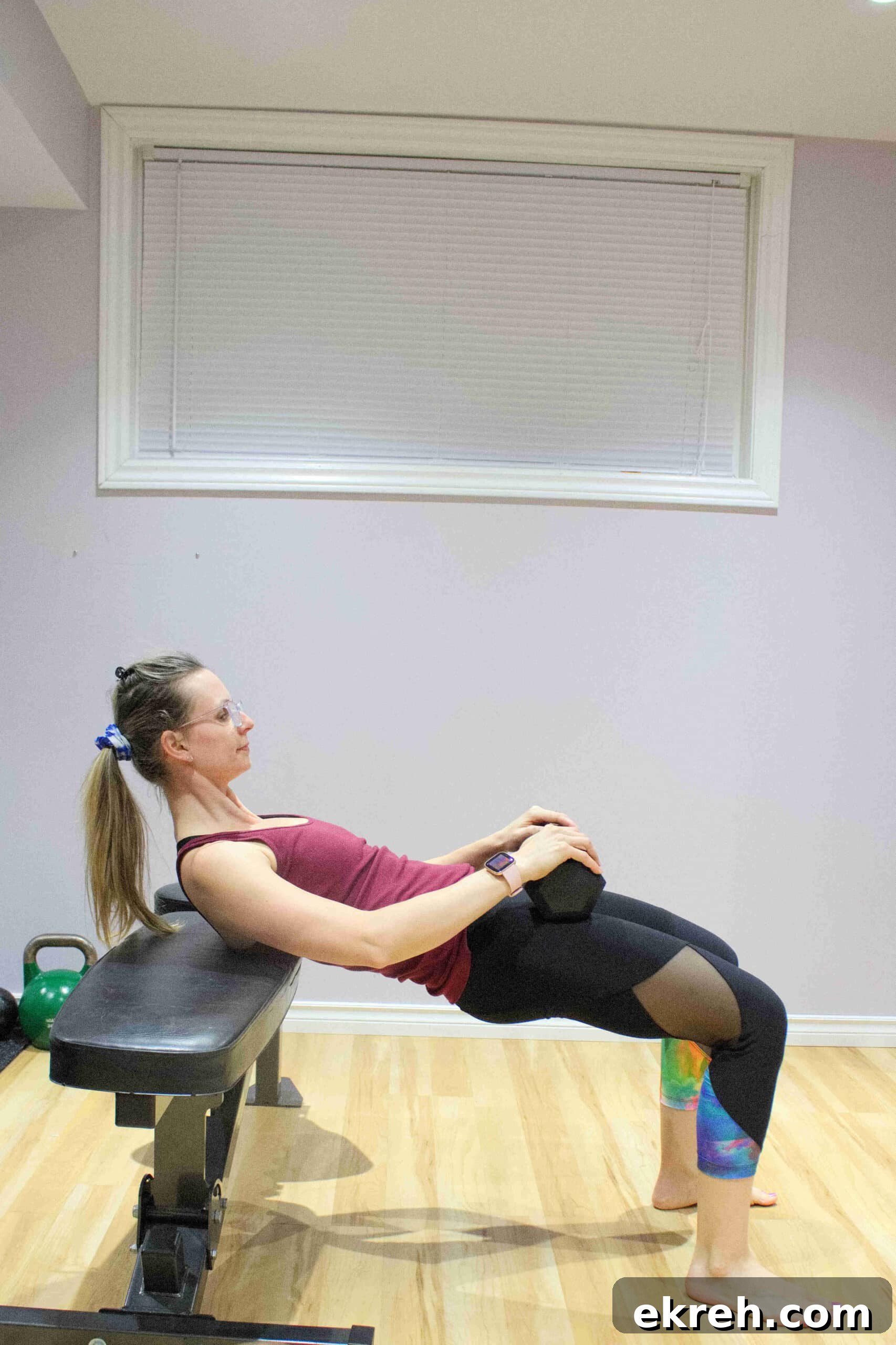 woman in pink shirt and dark pants performing a glute bridge on a bench with a dumbbell in her lap