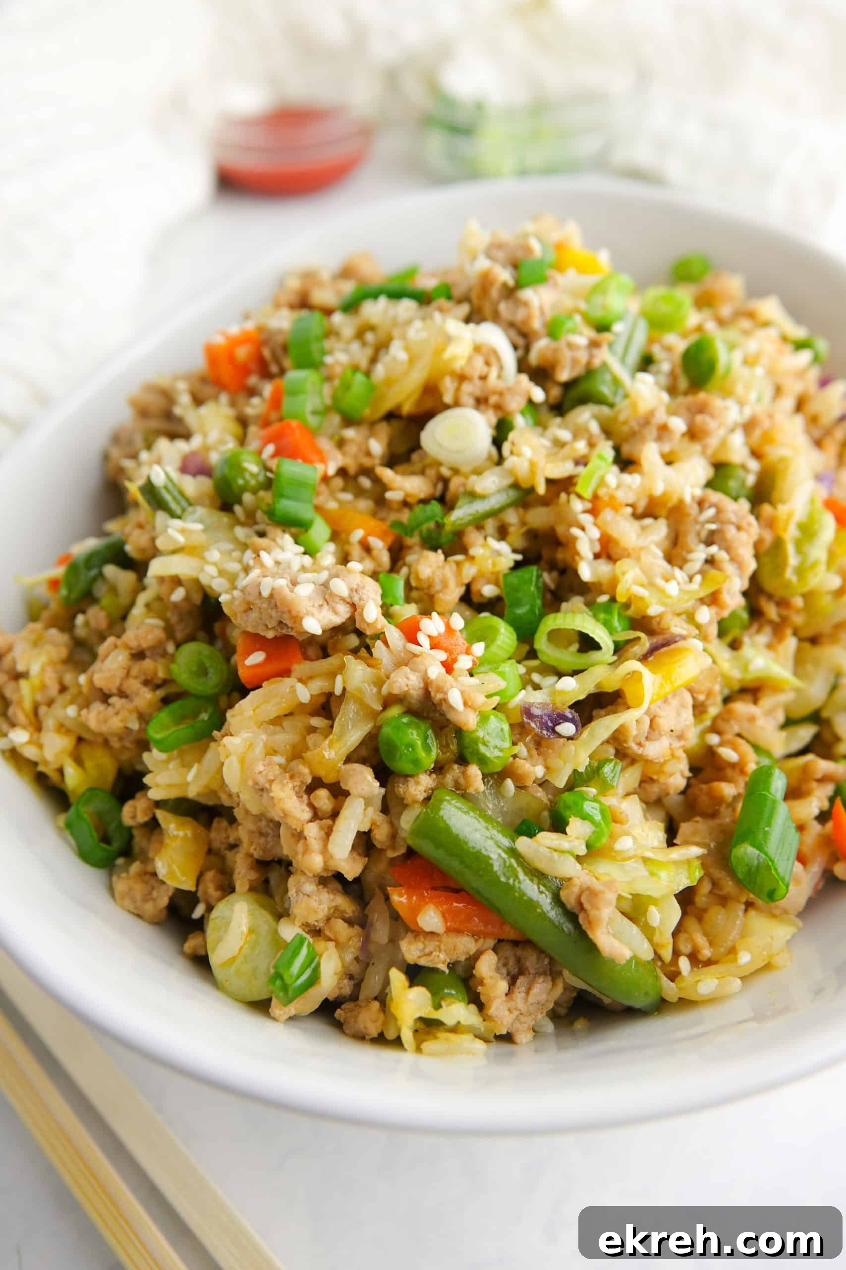 Unstuffed egg roll or pork fried rice presented in a white bowl on white marble countertop. Displayed with chop sticks, sriracha and green onions.