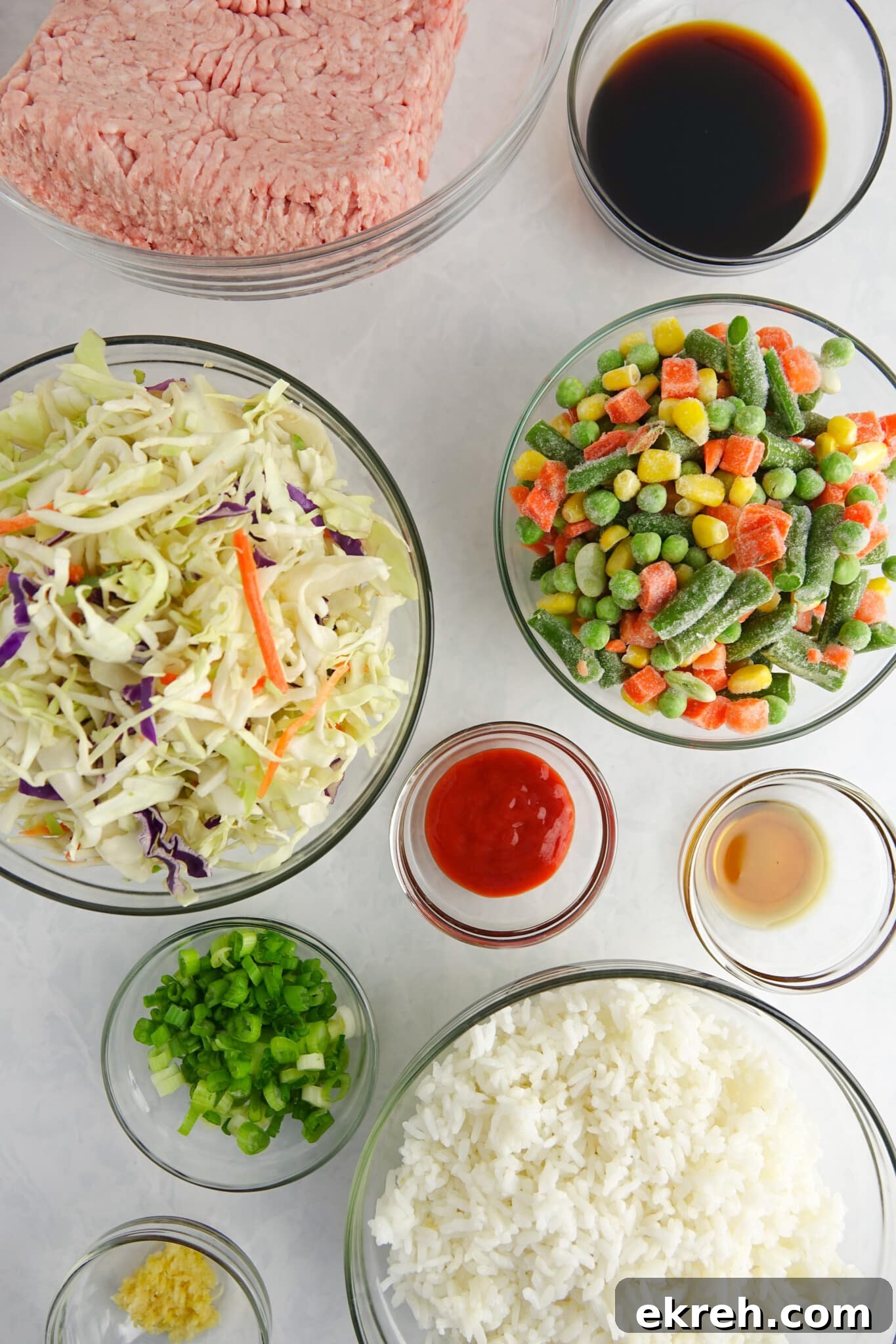 Ingredients for unstuffed egg rolls in a bowl displayed in clear glass bowls on a white marble counter: ground pork, soy sauce, coleslaw mix, frozen vegetables, sriracha, sesame oil, garlic, green onions and cooked rice.