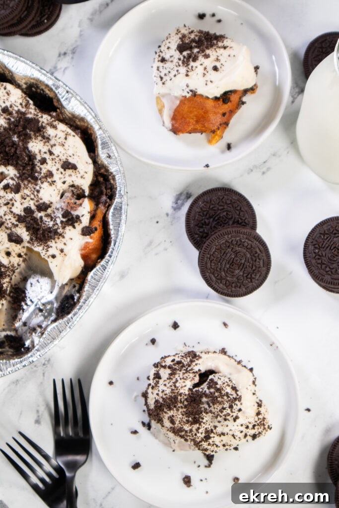 Air Fryer Oreo Cinnamon Rolls with Crunchy Oreo Swirl 3 Overhead view of an air fryer oreo cinnamon roll on a small white plate with another plate with a cinnamon roll, the remaining cinnamon rolls in the metal pan, and Oreo cookies scattered around.