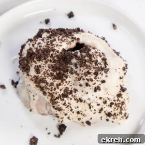 Air Fryer Oreo Cinnamon Rolls with Crunchy Oreo Swirl 6 An Air Fryer Oreo Cinnamon roll on a small white plate topped with cream cheese frosting and crushed Oreo pieces.