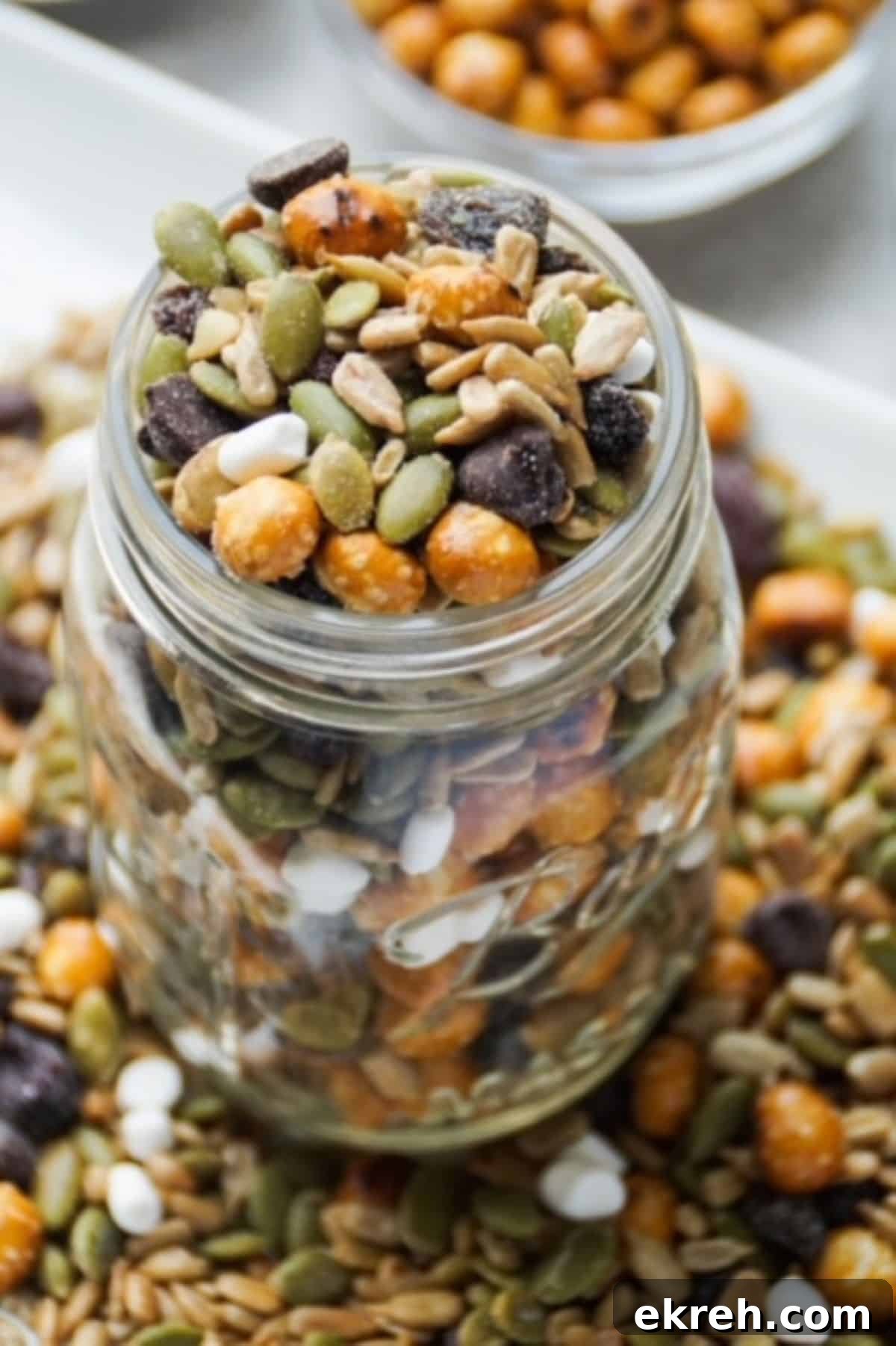 Nut-Free Trail Mix Recipe Kids Will Love 2 One glass mason jar filled with peanut-free trail mix. Base of jar surrounded by trail mix on a white plate. White bowl with pretzel balls partially pictured in the background.