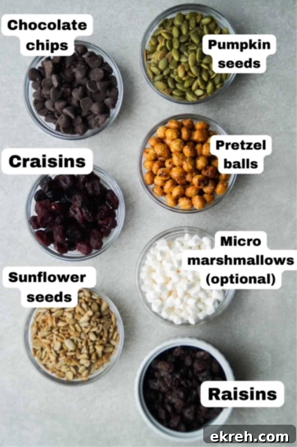 Nut-Free Trail Mix Recipe Kids Will Love 3 Trail mix ingredients in individual small glass bowls lined up into rows, each are labeled: Chocolate chips, craisins, sunflower seeds, pumpkin seeds, pretzel balls, micro marshmallows, and raisins.