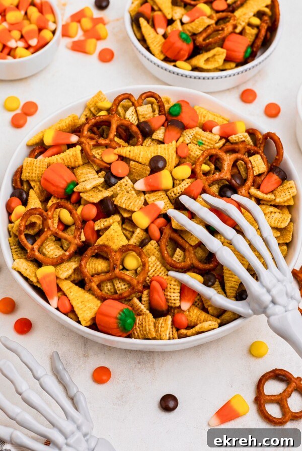 Large white bowl filled with Chex mix, pretzels, candy corn, and candies, with some scattered around the table and a scooper skeleton hand on top of the bowl.