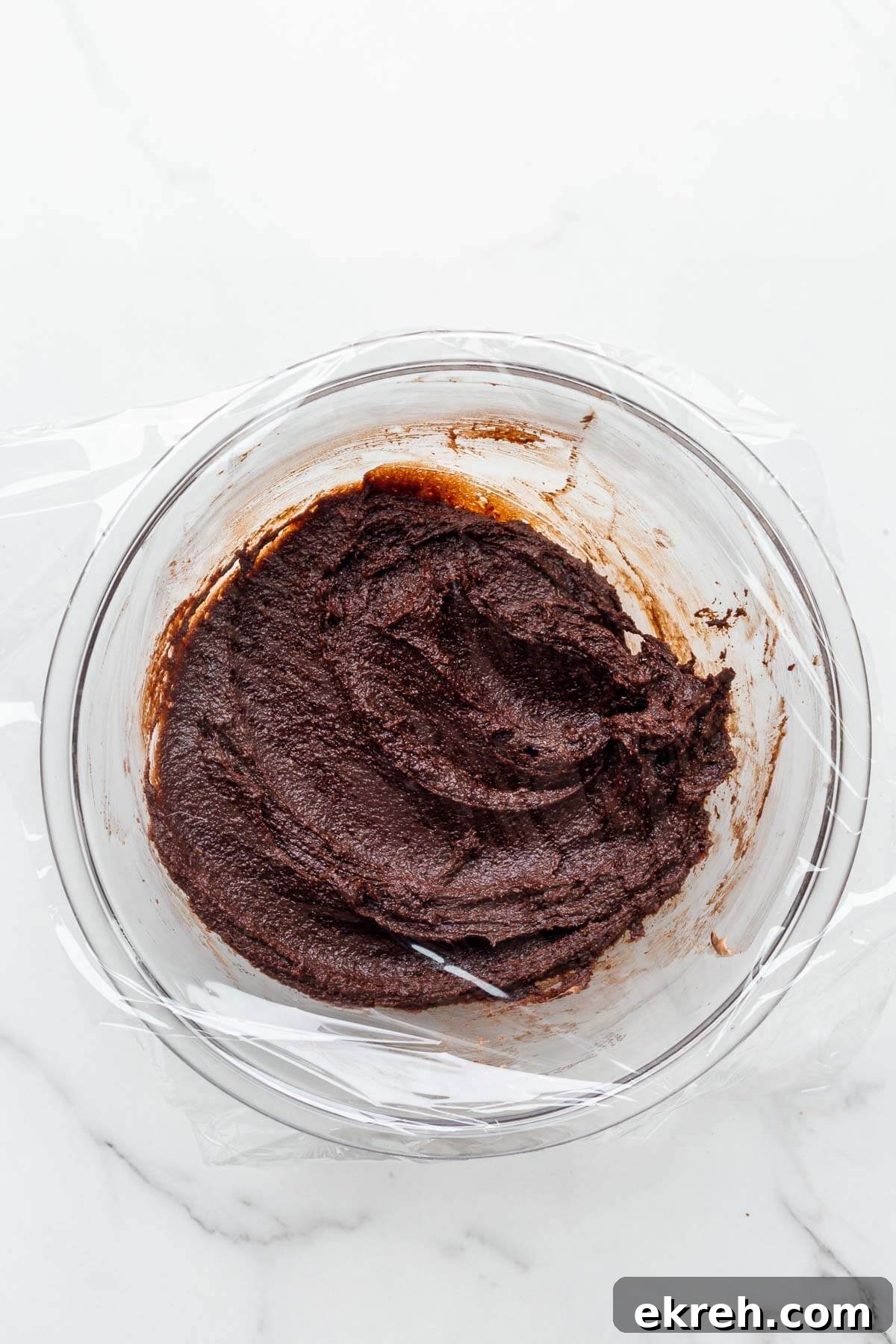 Chocolate financiers batter in a glass bowl wrapped in plastic wrap to chill it before baking.