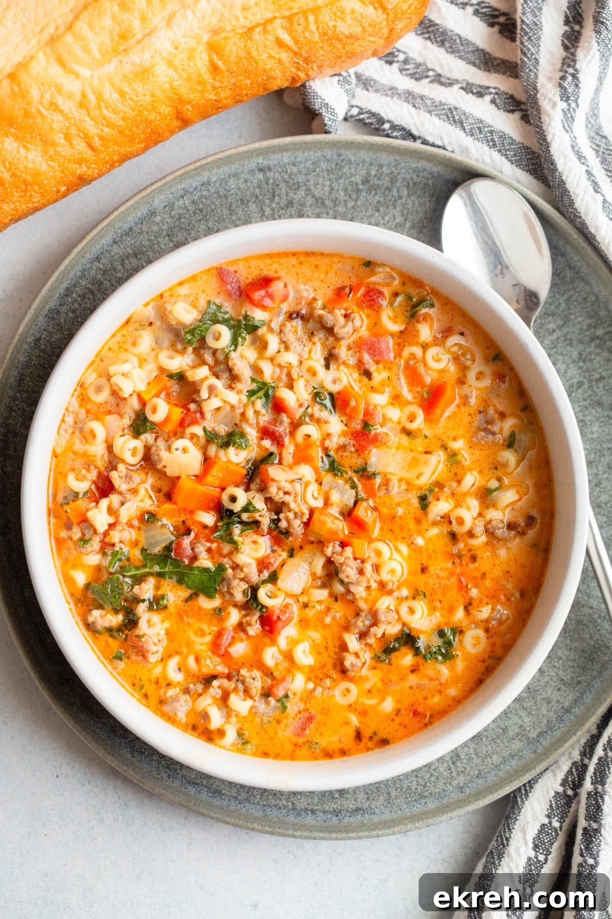 Creamy Italian Sausage Pasta Soup in a shallow cream colored bowl on a dark gray plate with a loaf of bread, a striped napkin next and spoon to it. 