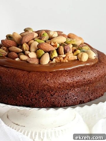 Caramel and nut topped chocolate cake on a white cake stand with white background.