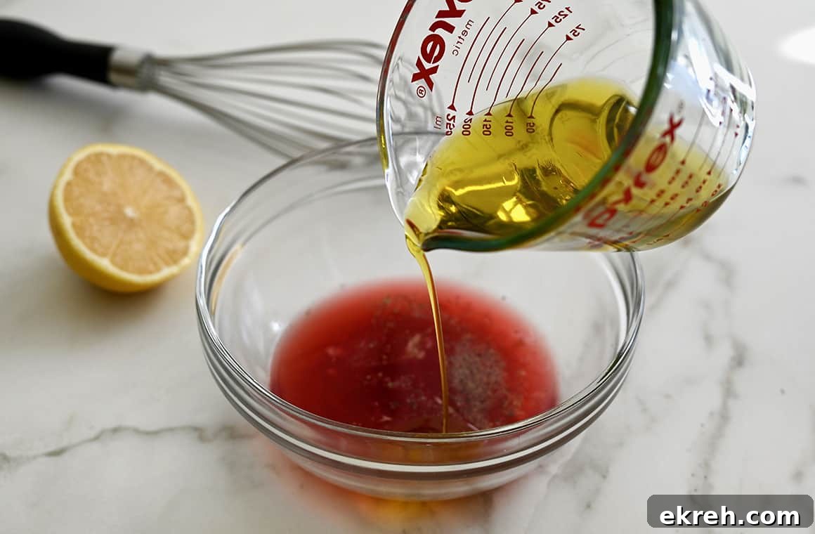 Olive oil in a glass measuring cup being poured into a clear bowl containing red wine vinegar and fresh lemon juice