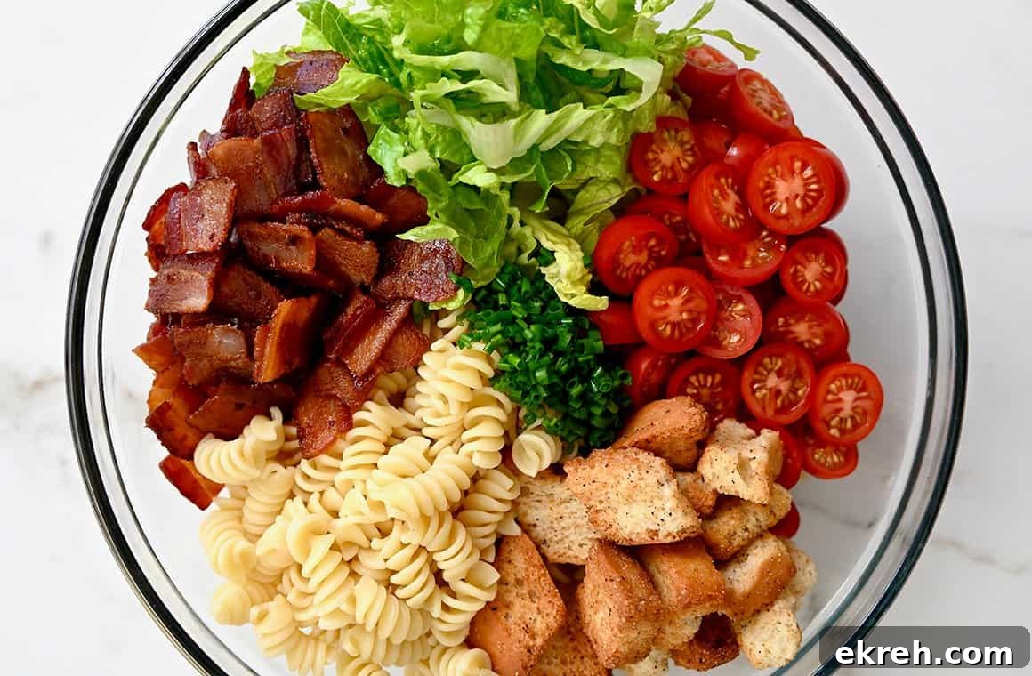 A top-down view of a clear bowl containing chopped bacon, lettuce, halved cherry tomatoes, croutons and rotini
