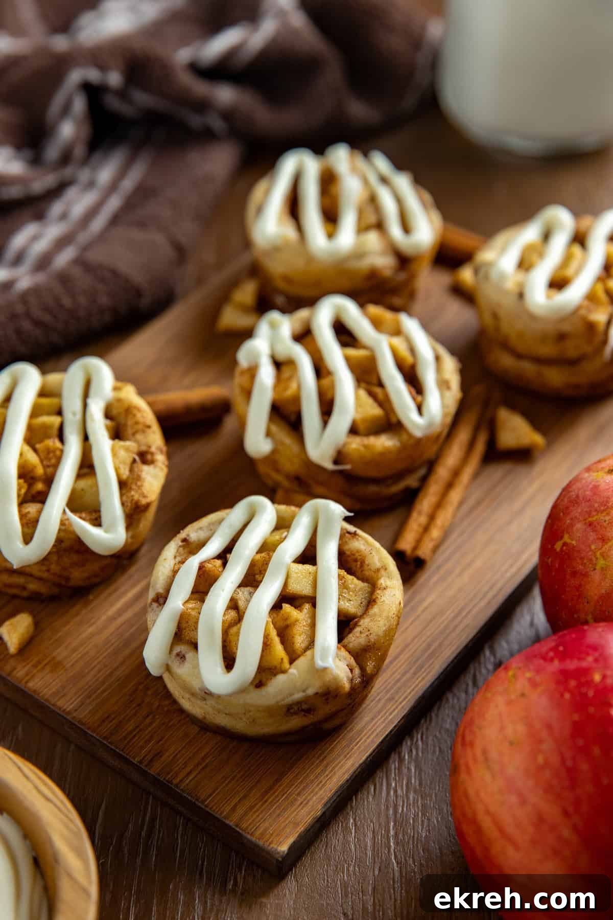 Apple Pie Filled Cinnamon Rolls with Caramel Glaze 2 Five baked and frosted apple pie cinnamon rolls on wooden cutting board with some cinnamon sticks and fresh red apples on the side as garnish.