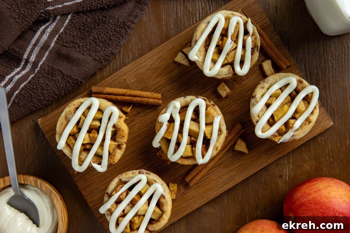 Apple Pie Filled Cinnamon Rolls with Caramel Glaze 9 Overhead view of five baked and frosted apple pie cinnamon rolls on wooden cutting board with some cinnamon sticks and fresh red apples on the side as garnish.
