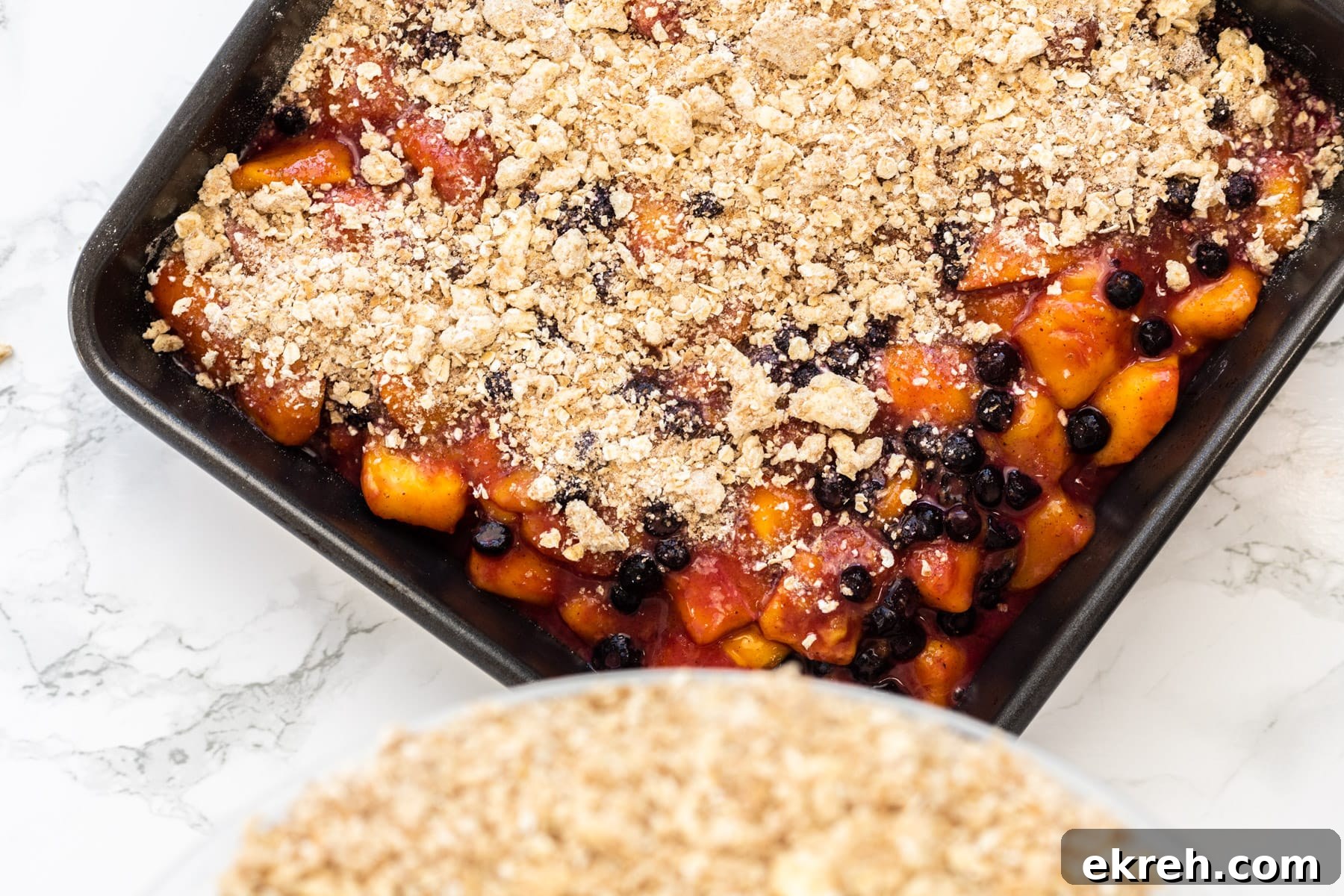 Pouring crumble topping over a baking dish filled with spiced mangoes and blueberries.