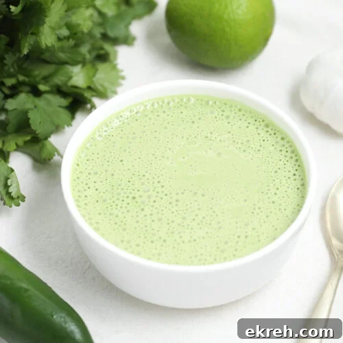 A white bowl filled with creamy green sauce sits on a white surface, surrounded by cilantro, a lime, garlic, a jalapeño, and a gold spoon.