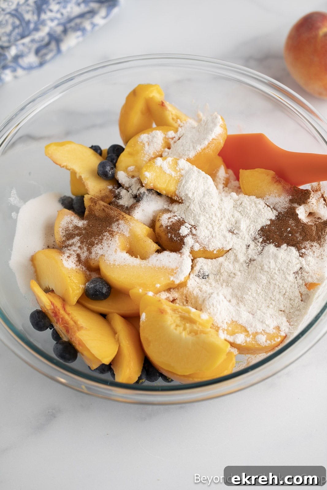 Mixing sliced peaches, blueberries, flour, and spices together in a large glass bowl. 