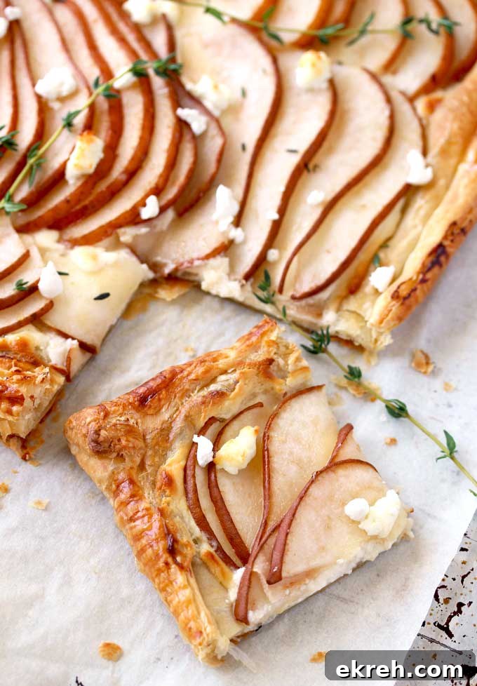 View of a golden brown puff pastry tart sliced The tart is topped with thinly sliced pears garnished with goat cheese and fresh thyme on a parchment paper lined baking sheet.
