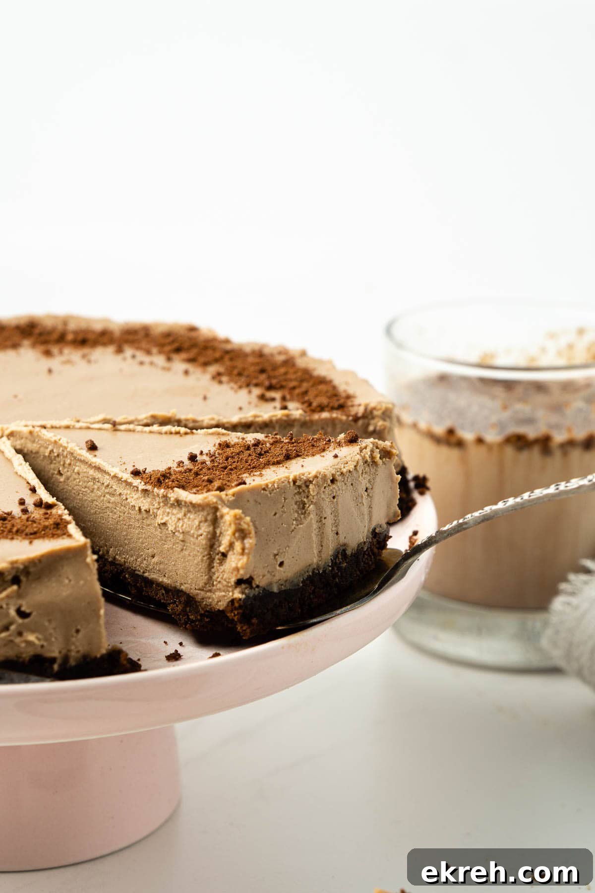 Creamy No-Bake Vegan Milo Cheesecake Recipe 4 Slice of milo cheesecake being removed from a pink cake stand with a glass of Milo in the background.
