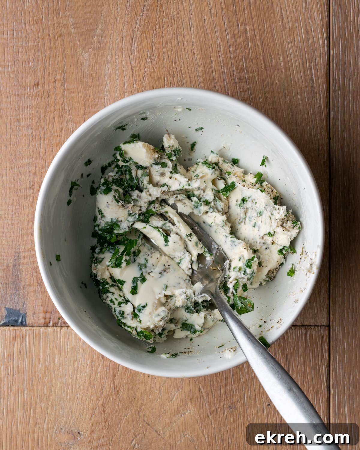 A fork is used to mix herbs and seasonings into a bowl of softened butter.