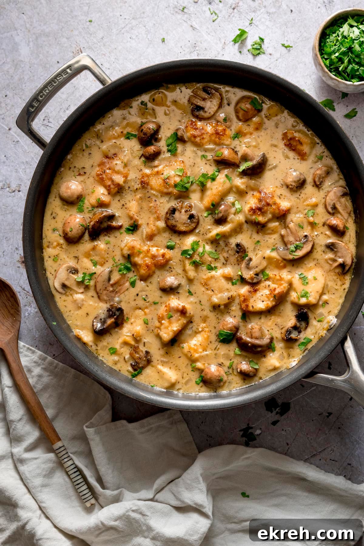 Overhead image of creamy chicken and mushroom casserole in a large frying pan on a light background. There is a cream linen napkin and a wooden spoon next to the pan and parsley scattered in and around the pan.