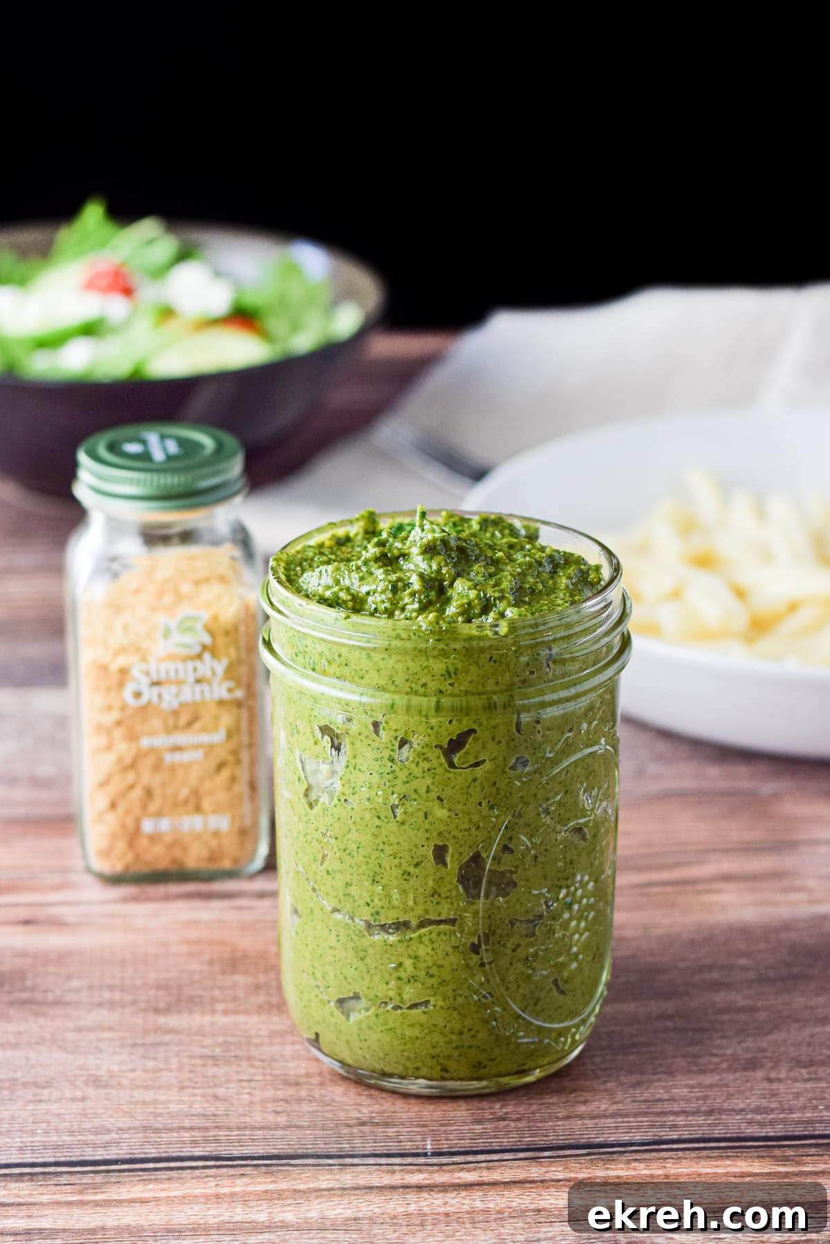 A jar with pesto in it, with nutritional yeast, pasta, and salad in the back